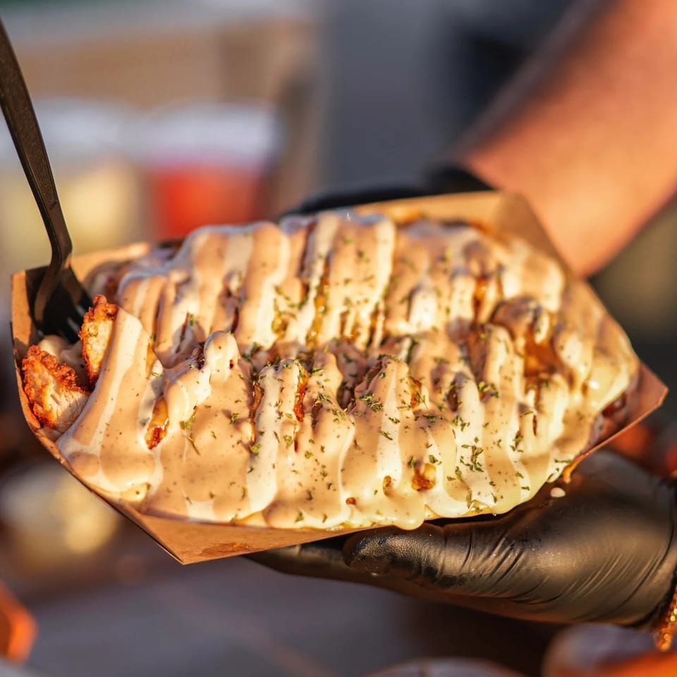 Hands in black gloves holding a tray of Hot Chicks Food Truck's loaded fries with sauce and herbs.
