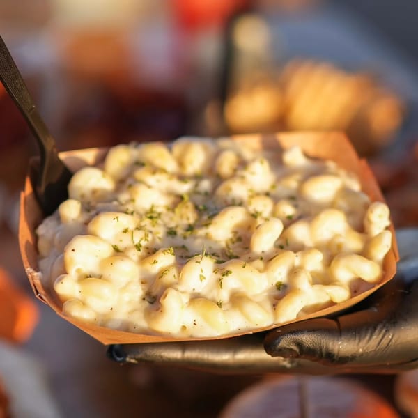 Creamy mac and cheese with herbs in a paper tray, held by a gloved hand.