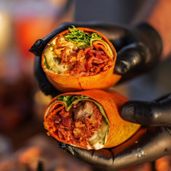 Gloved hands holding two halves of a Hot Chicks Food Truck chicken wrap with crispy bacon and lettuce.