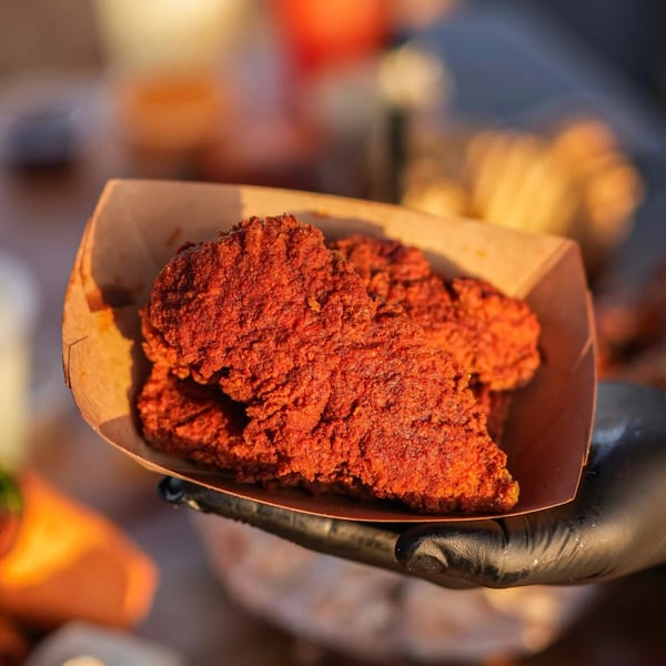 A hand in a black glove holds a cardboard tray filled with crispy, red-spiced Hot Chicks chicken tenders.