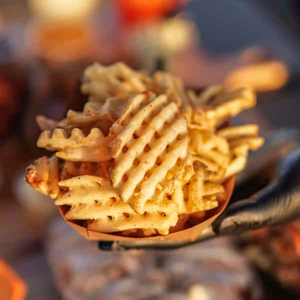 A hand in a black glove holds a paper tray filled with crispy waffle fries from Hot Chicks Food Truck.