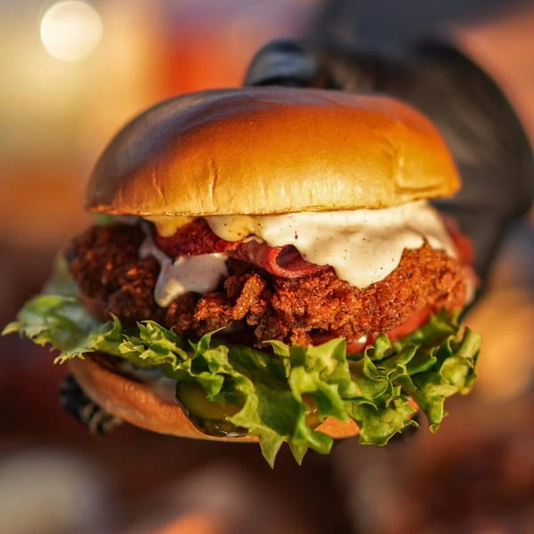A hand in a black glove holds a Hot Chicks Food Truck fried chicken sandwich with sauce, bacon, lettuce, and pickles.